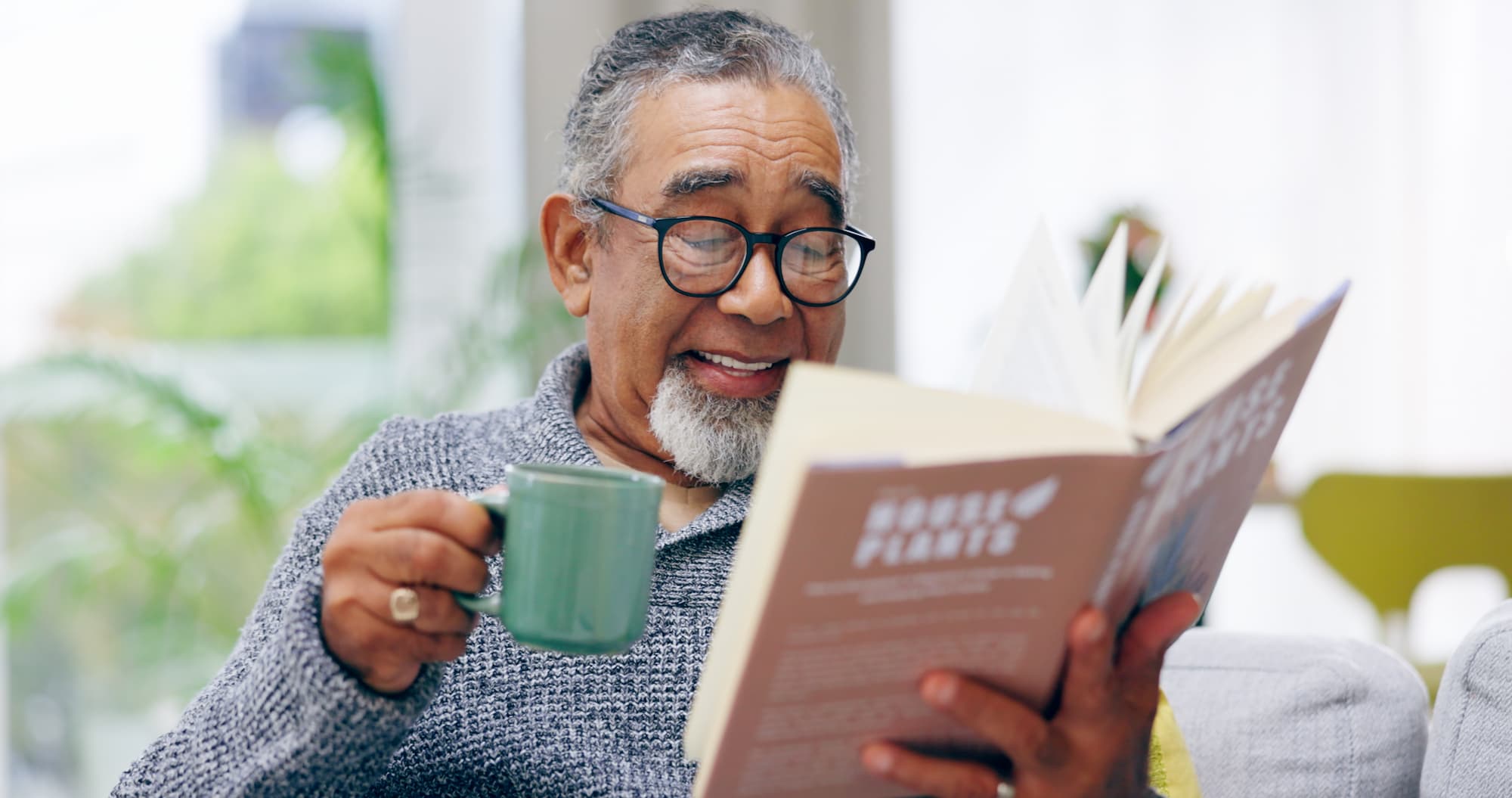 A senior man with a green coffee cup in hand laughing while reading a book from the comfort of his home and chair