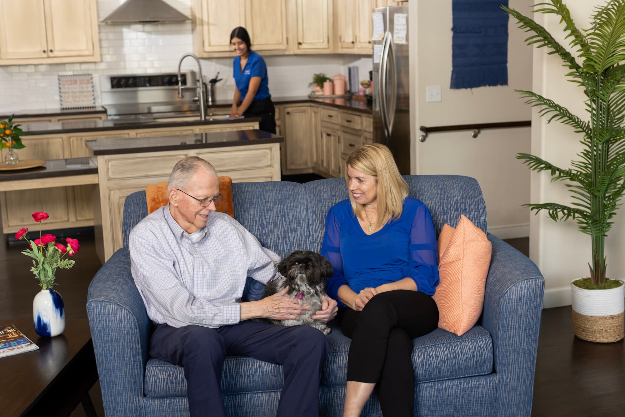 A woman staff member and elderly male on a blue couch smiling together with a dog sitting in between them